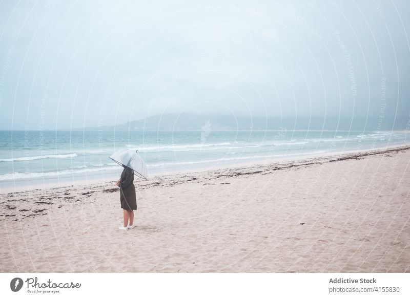 Anonymous person with umbrella walking on sandy beach sea alone travel ocean lonely stormy overcast traveler shore coast nature wave weather tourism seaside