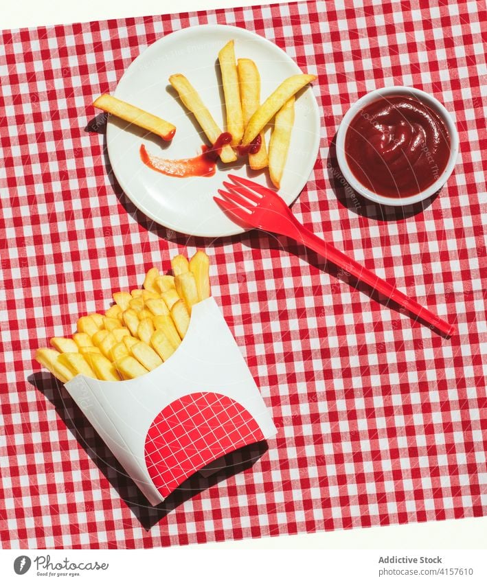 French fries packet near plate with potatoes soaked in ketchup american culture condiment potato chips unhealthy eating red background white frites take out