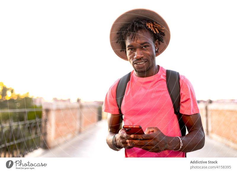 Smiling ethnic man on bridge in summer traveler smartphone tourist handsome using city sightseeing male black african american backpack casual trip message