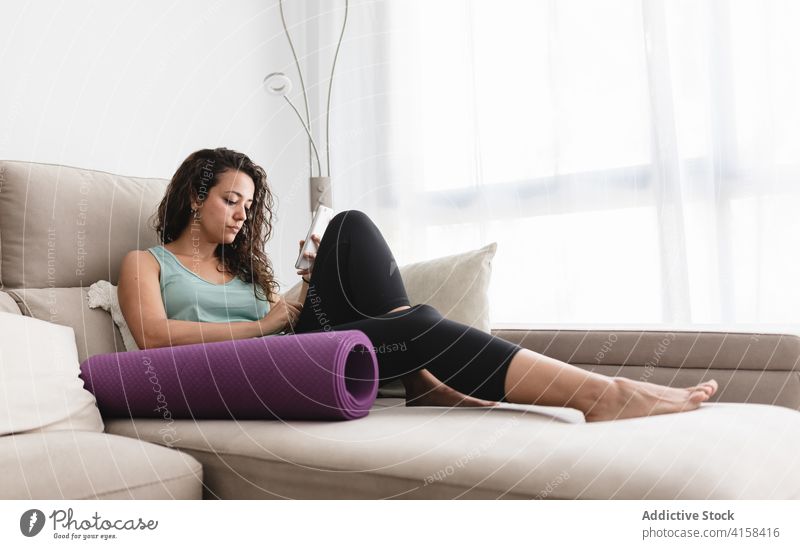 Tranquil woman relaxing on sofa with smartphone and yoga mat browsing home lying using weekend female sportswear activewear living room couch soft modern