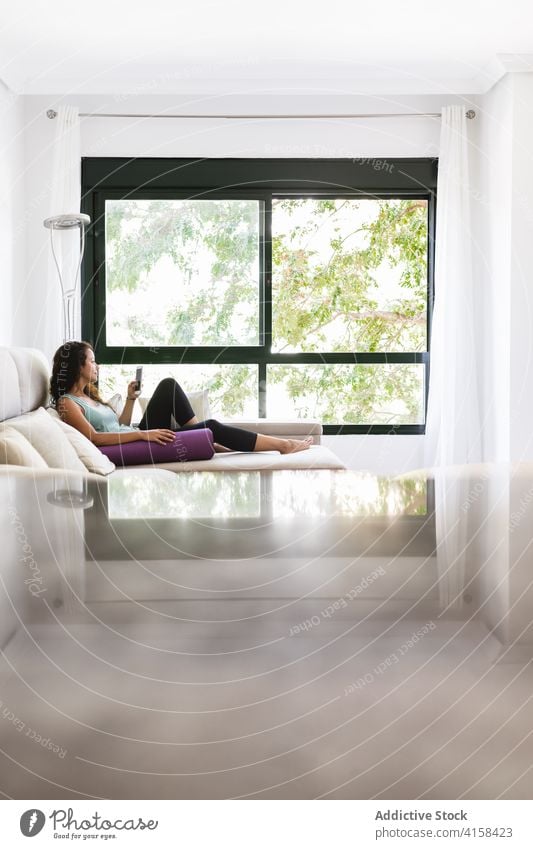 Tranquil woman relaxing on sofa with smartphone and yoga mat browsing home lying using weekend female sportswear activewear living room couch soft modern