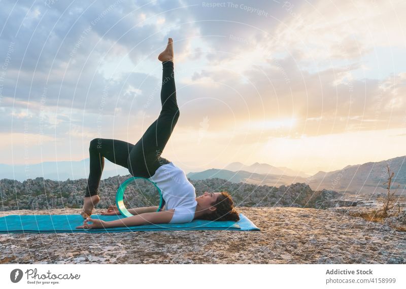 Flexible woman practicing asana with yoga wheel backbend bridge variation stretch flexible equipment eka pada setu bandha sarvangasana nature pose mountain rock