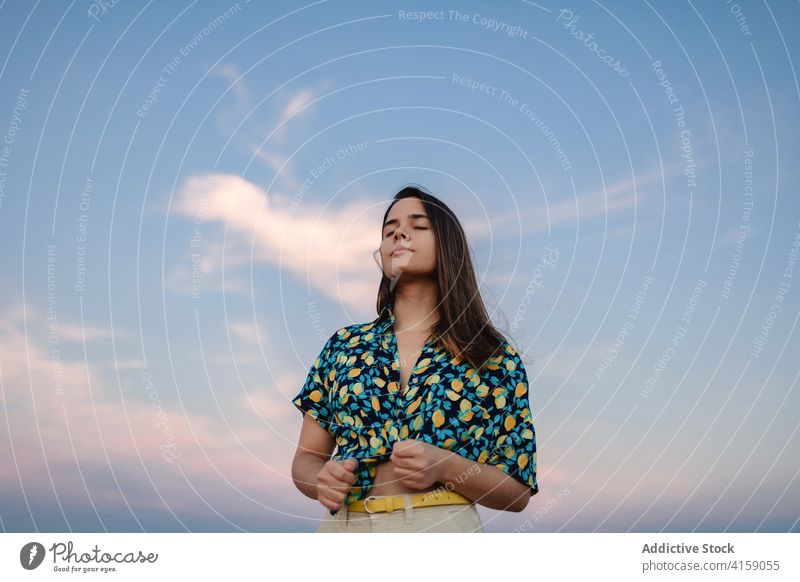 Dreamy woman under cloudy sky at sunset eyes closed dreamy blue sky ornament style harmony idyllic appearance trendy apparel charming feminine stylish wear