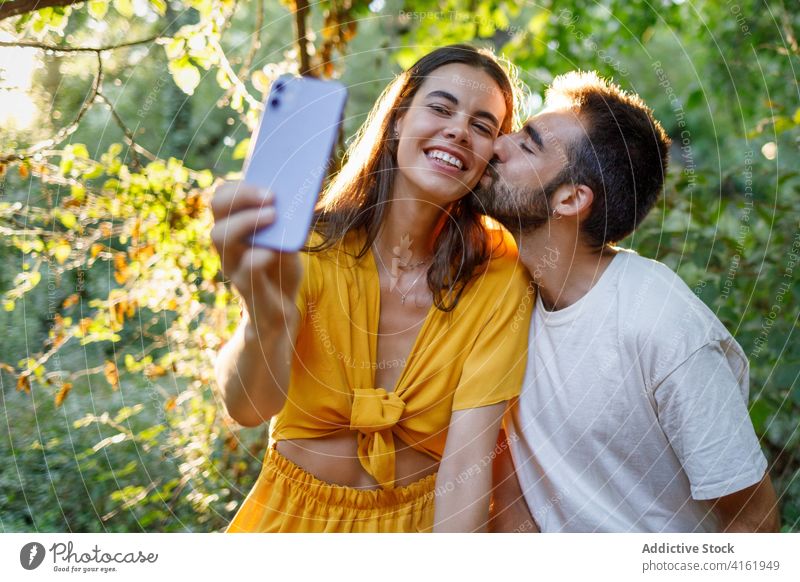 Cheerful ethnic couple taking selfie in park kiss happy smartphone cuddle date romantic love together relationship gadget mobile girlfriend boy young casual