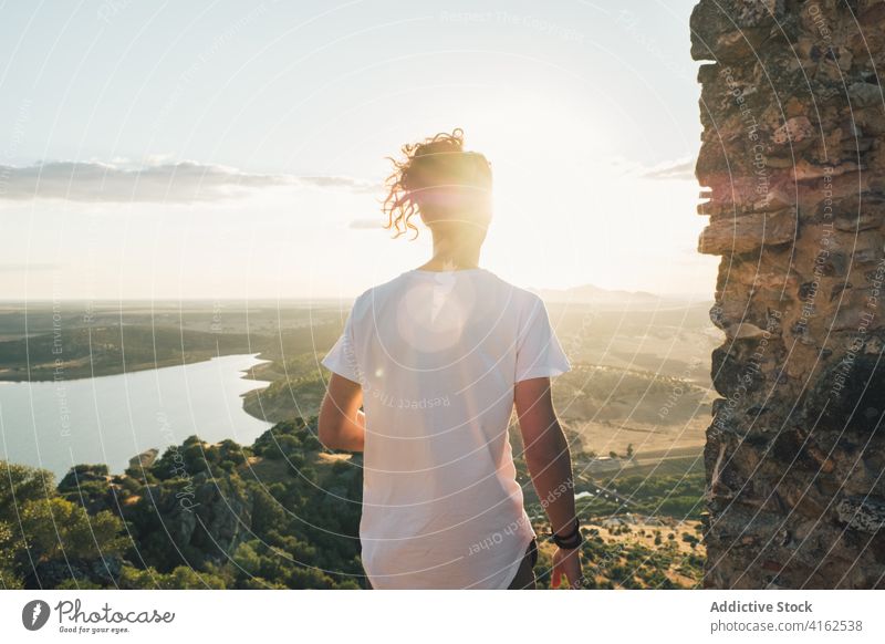 Unrecognizable man standing on hill above sunny valley picturesque admire nature pond greenery scenic viewpoint landscape hilltop wanderlust travel amazing