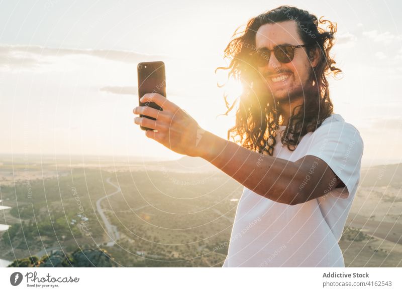 Cheerful man taking selfie against sunny lush valley traveler cheerful toothy smile smartphone nature excited moment gadget hilltop wanderlust amazing viewpoint