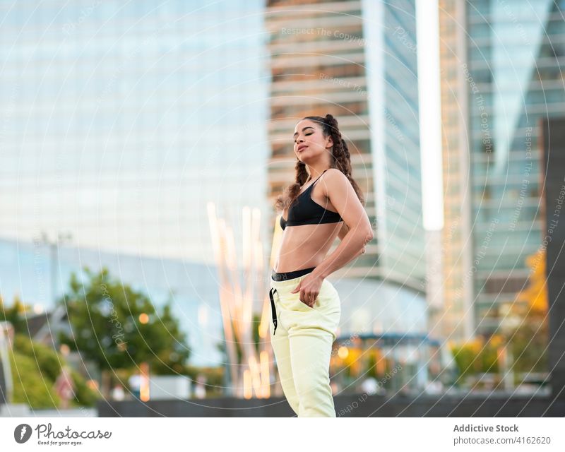 Confident young female athlete resting in park near stylish city buildings woman self assured hand in pocket skyscraper recreation sportswoman urban healthy