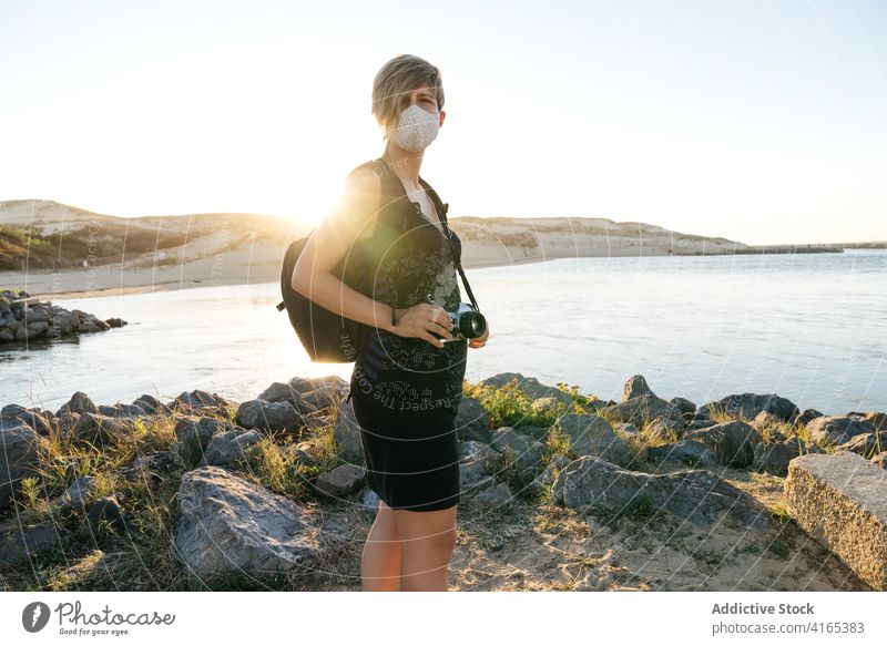 Woman admiring sea view from rocky coast at sunset woman seashore traveler vacation journey new normal trip holiday relax female short hair backpack mask