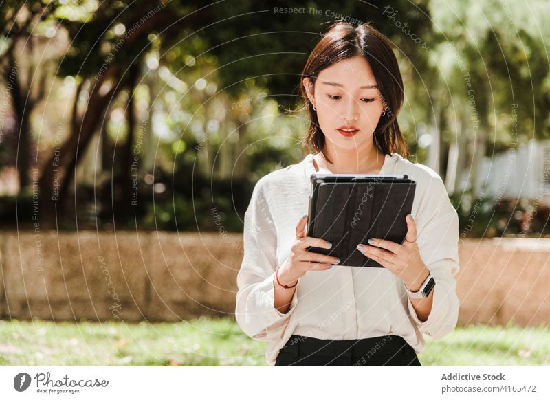 Young ethnic woman working remotely on tablet in park attentive new normal coronavirus job busy gadget using browsing female young asian formal mask covid 19