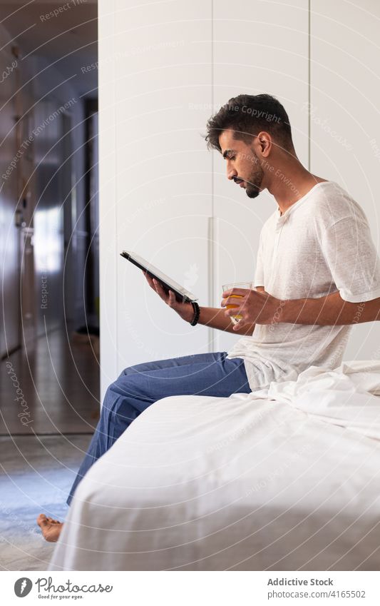 Young man using tablet in bedroom morning drink gadget at home device young beard browsing internet read online lifestyle connection surfing beverage modern