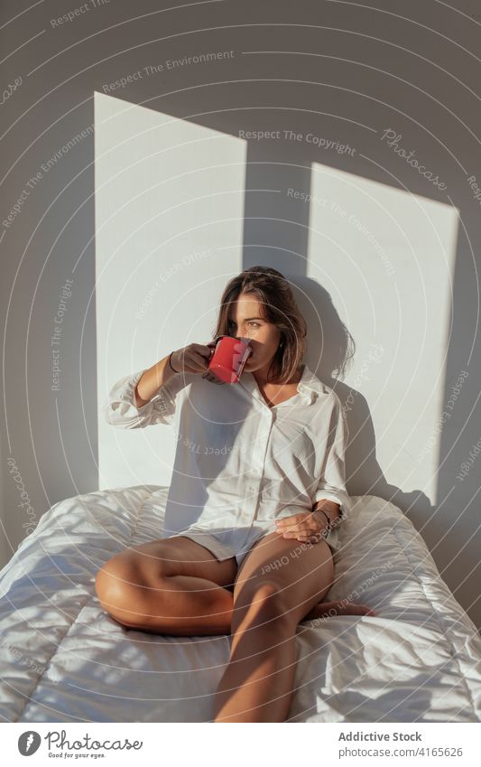 Cheerful young woman drinking coffee in bed morning smile happy sensual comfort allure cozy joy female white shirt body barefoot home beverage sunny relax rest