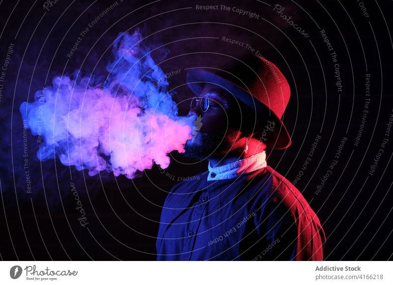 Ethnic man smoking in studio with neon lights smoke vape steam style illuminate dark male ethnic black african american sunglasses hat trendy hipster cool