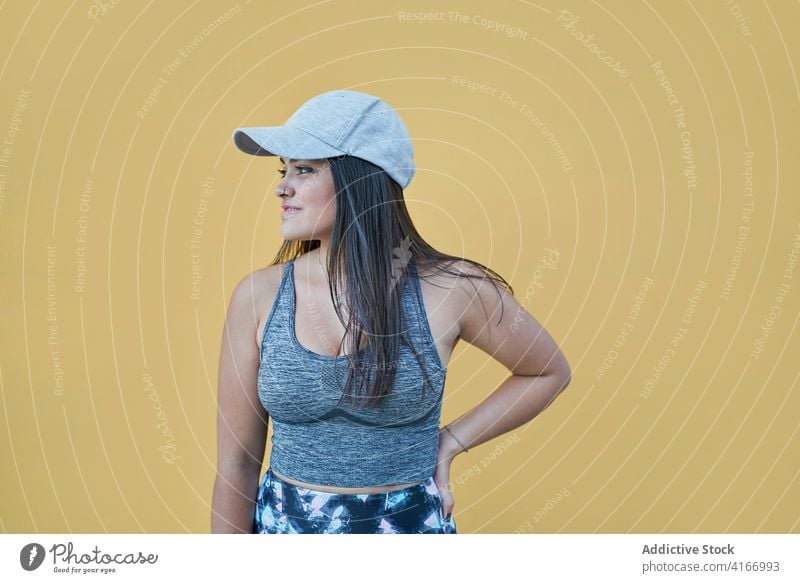 Cheerful woman with hand on waist standing against yellow wall toothy smile cheerful happy charismatic optimist joy cool expressive energy cap young confident