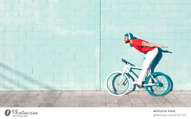 Young man riding bicycle and listening to music in headphones on city street ride bike cyclist active trick trendy building stone wall male young beard outfit