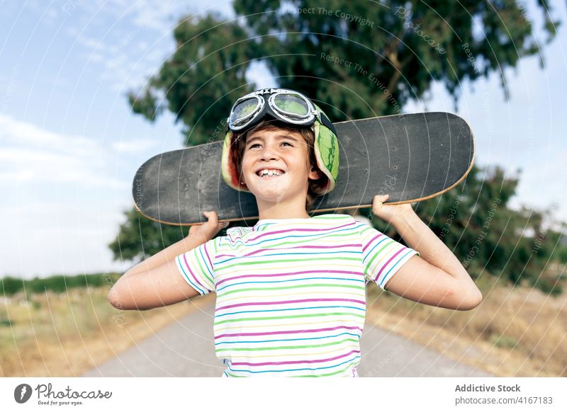 Happy dreamy boy with skateboard on roadway child goggles happy countryside stylish apparel cloudy blue sky childhood kid positive smile enjoy stripe wear