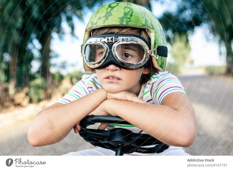 Crop melancholic trendy kid in goggles and watermelon head wear boy melancholy headwear dreamy steering wheel lean on hand portrait road swimming ponder