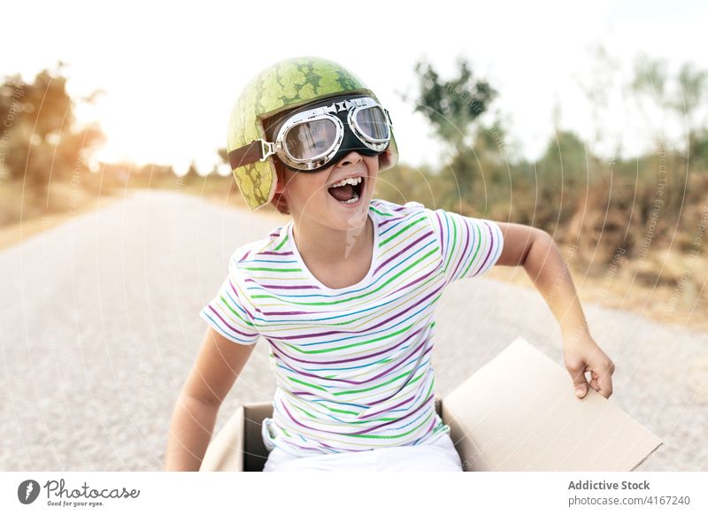 Excited boy in goggles sitting in cardboard box on road excited having fun childhood mouth opened stylish carefree sky kid positive smile enjoy stripe wear