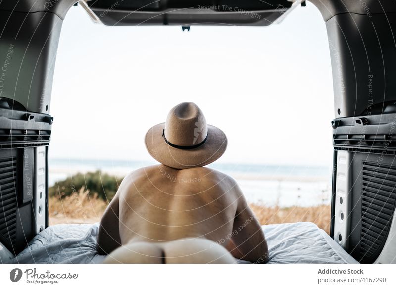 Unrecognizable naked man in hat resting on mattress in vehicle tourist contemplate ocean vacation sky lying horizon endless felt nude travel transport journey