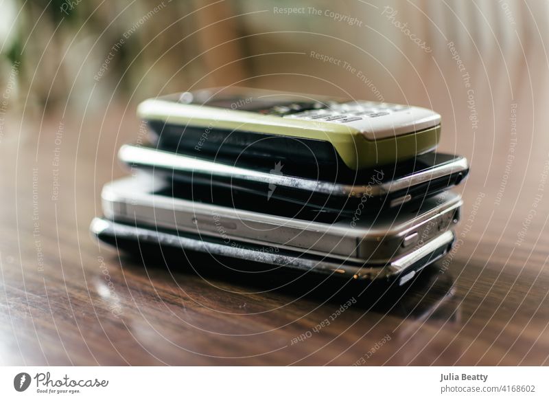 Stack of four old cellphones on a dining room table; top phone is green outdated cellular hold mobile wireless recycle e-cycle trash garbage collect collection