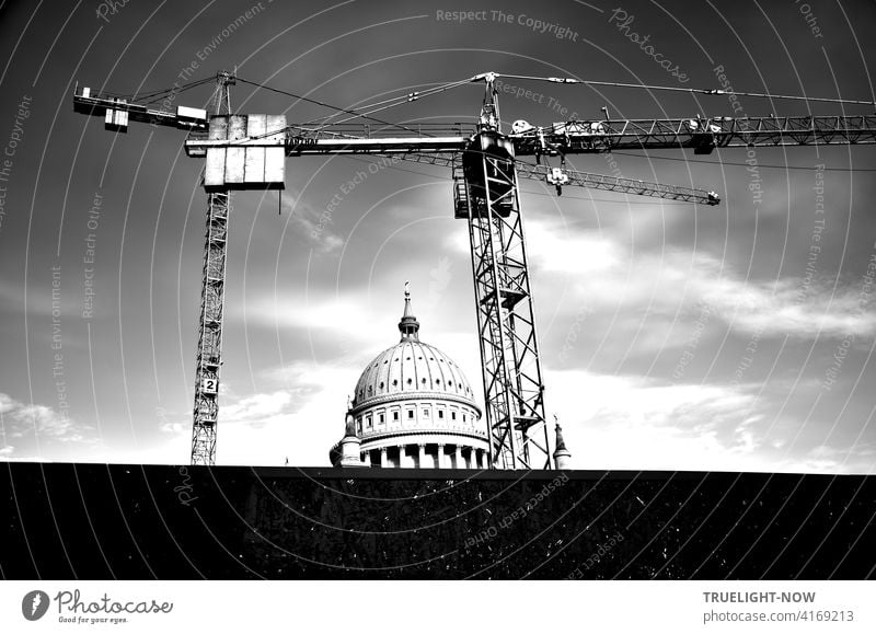 A construction crane and another construction crane behind a dark screen wall form the frame around the round tower of the Nikolai church, a landmark of Potsdam, in front of a slightly cloudy sky.