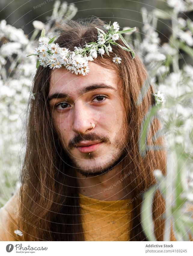 Young man with flowers in long hair sitting in field gay romantic bloom relax rest nature peaceful calm young male brunet lifestyle homosexual gender lgbt lgbtq