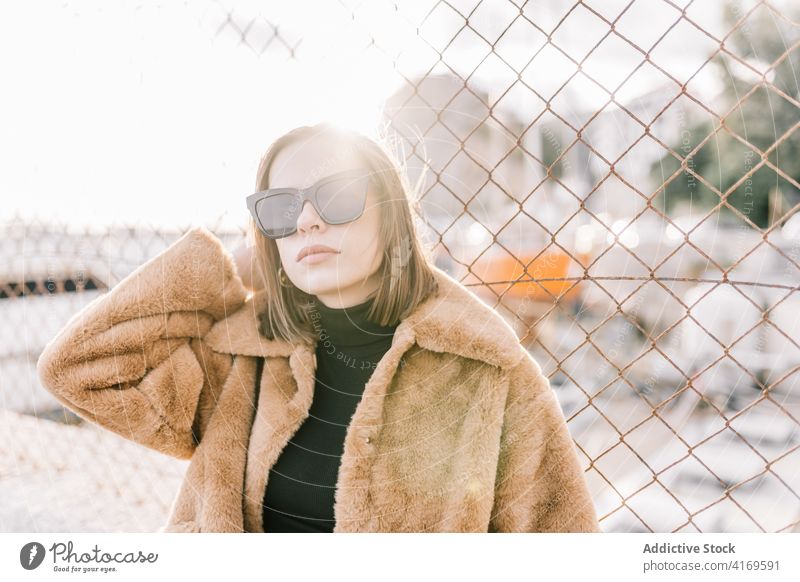 Stylish woman in warm jacket in city fur style trendy outfit determine sunglasses outerwear female net fence street urban cool fashion serious town calm