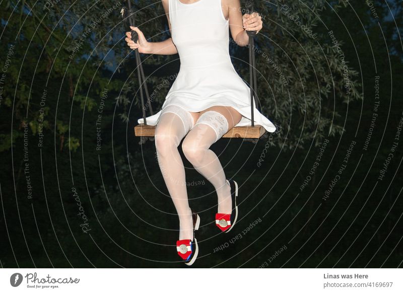 A woman dressed in a sexy white outfit with her extremely long legs swinging under the bridge. The sky is dark blue, the bushes are green, and the mood is fine. Her fashionable blue shoes are shining from far away.