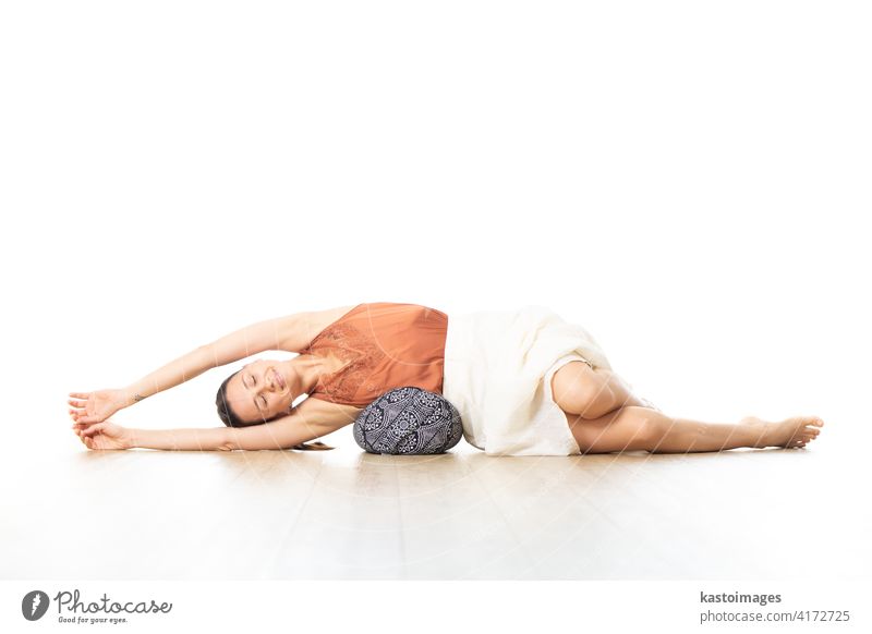 Restorative yoga with a bolster. Young sporty attractive woman in bright white yoga studio, lying on bolster cushion, stretching and relaxing during restorative yoga. Healthy active lifestyle