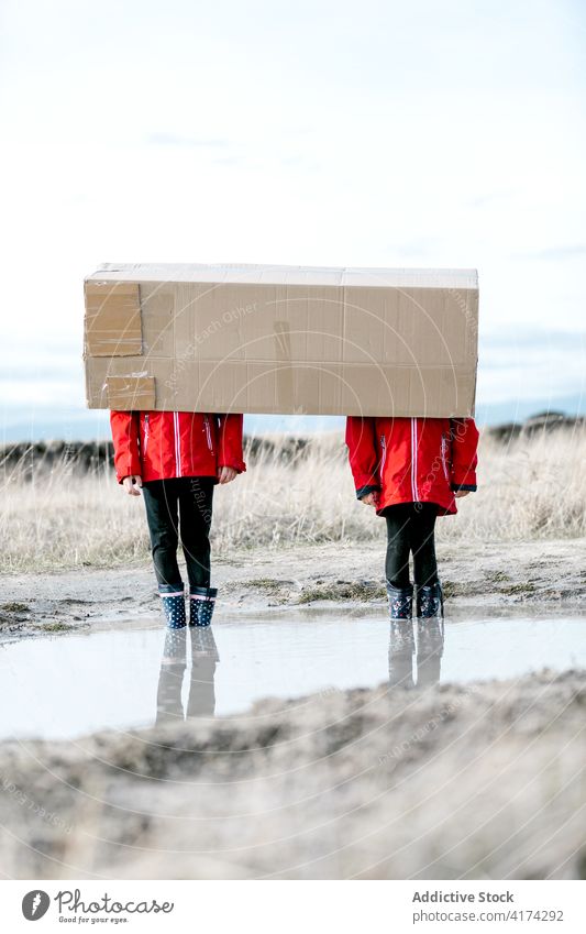 Unrecognizable kids playing with carton package children cardboard having fun hide cover face together puddle rubber boot raincoat playful childhood friendship