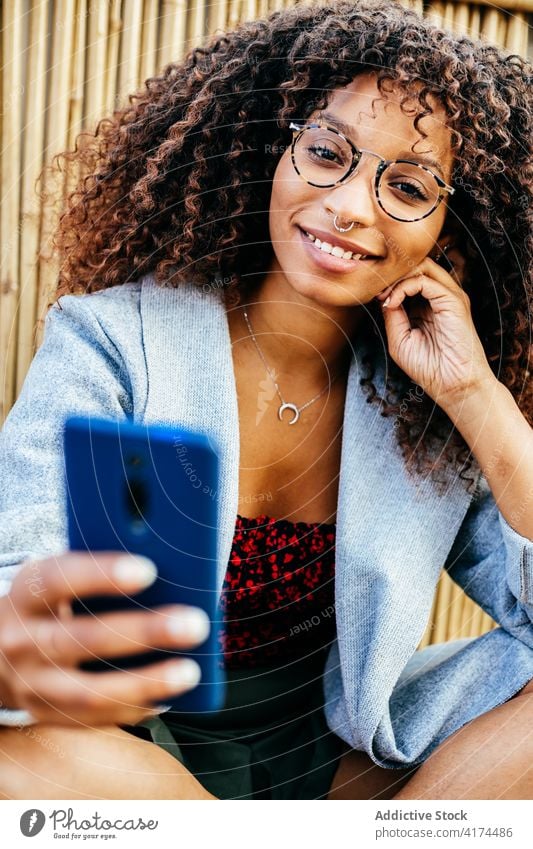 Cheerful black woman using smartphone near bamboo wall rest style floor wooden smile legs crossed female young outfit weekend trendy ethnic african american