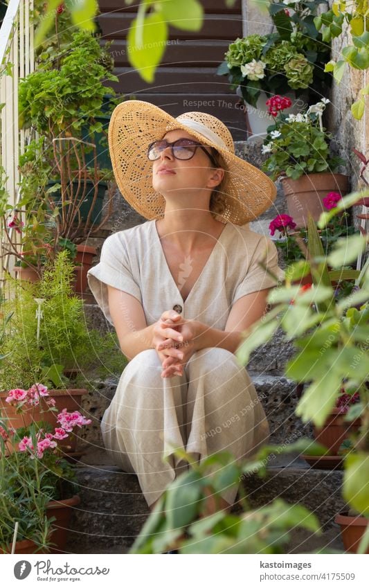 Beautiful female tourist wearing big straw sun hat and sundress sitting and relaxing on old stone house stairs during summer travel on Mediterranean cost on hot summer day