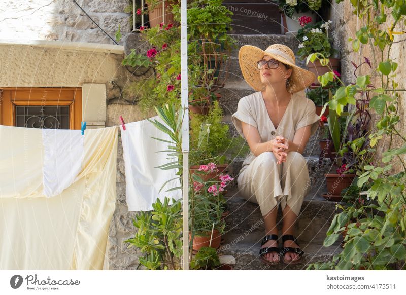 Beautiful female tourist wearing big straw sun hat and sundress sitting and relaxing on old stone house stairs during summer travel on Mediterranean cost on hot summer day