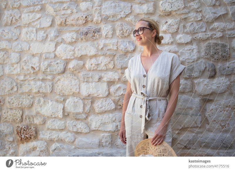 Portrait of beautiful cheerful blonde woman wearing one piece sundress and summer hat, standing in front of old medieval stone wall. Summer vacation portrait concept. Copy space.