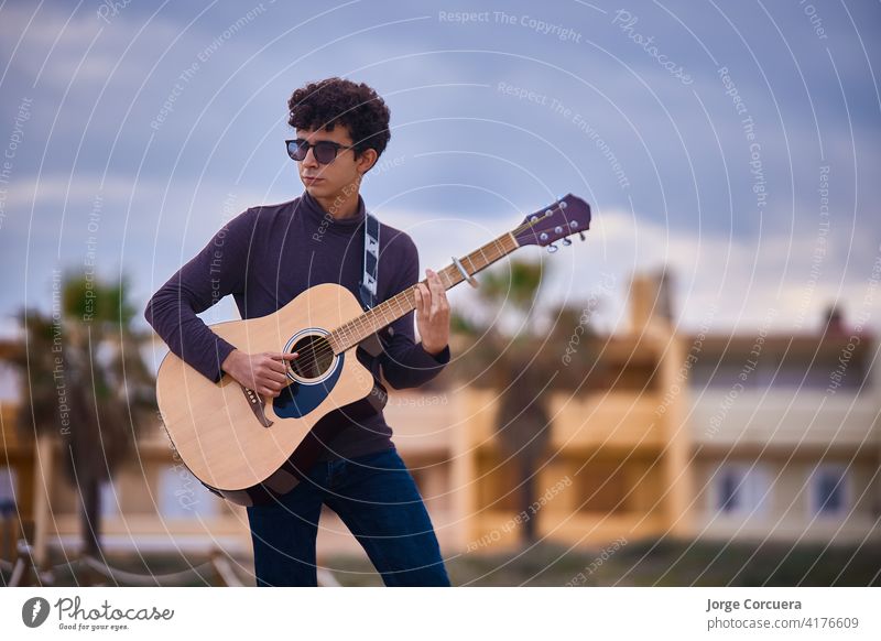 young caucasian teenage boy playing acoustic guitar on the beach. sunglasses and dark clothing. art artist blues casual composer concert entertainment