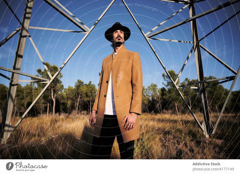 Stylish man in hat and coat standing near metal tower style trendy confident modern serious young beard male ethnic lifestyle outfit personality fashion pensive