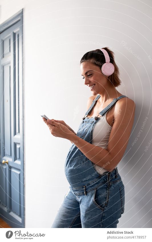 Pregnant woman enjoying music in headphones at home pregnant listen pregnancy overall song wireless female smile cheerful room melody sound playlist happy tune