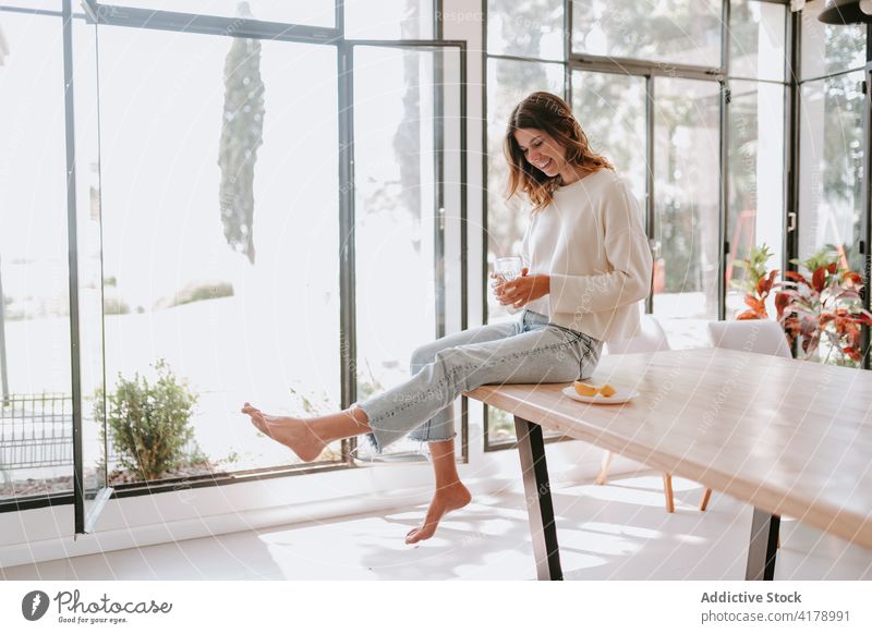 Content woman with glass of water with lemon in kitchen healthy drink diet morning cheerful home female table carefree sit young enjoy fresh relax refreshment