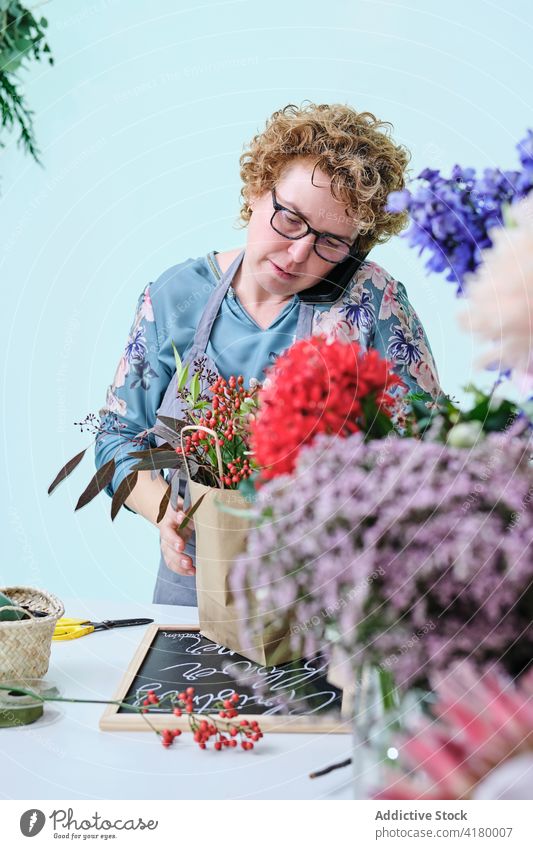 Woman making composition of flowers in shop florist woman bouquet arrangement smartphone talk multitask floristry female craft paper bag retail work plant