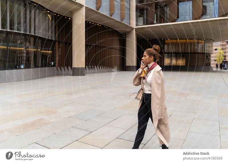 Focused young African American female strolling in city and talking on smartphone woman walk phone call square conversation busy pedestrian style speak