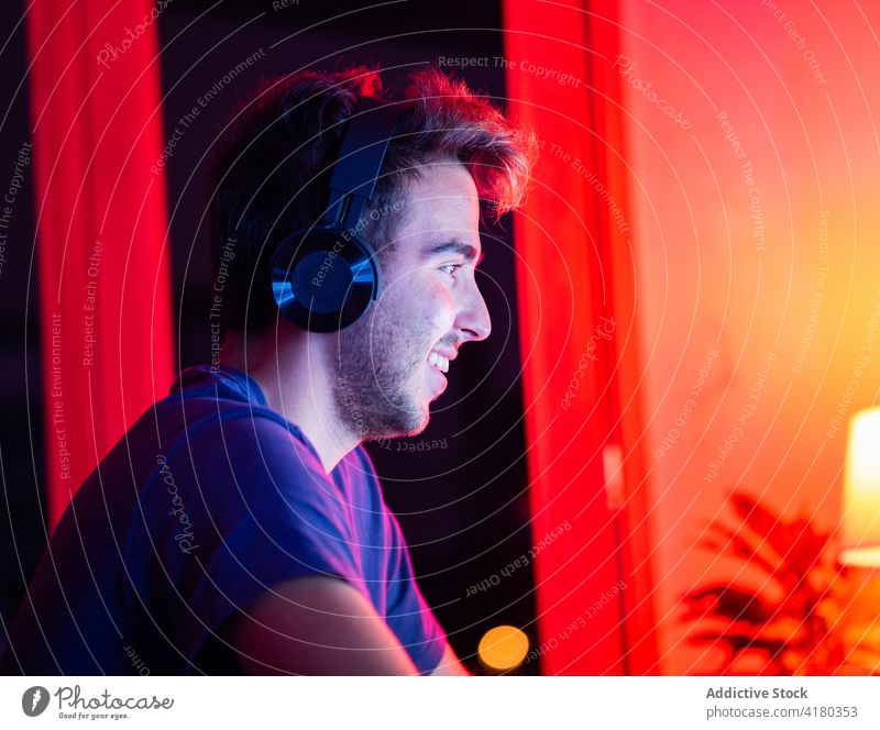 Cheerful man in headset sitting at desk with wireless headphones happy toothy smile lean on cheerful work gadget dark using computer handsome casual device