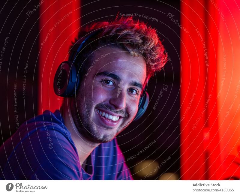 Cheerful man in headset sitting at desk with computer happy toothy smile lean on cheerful headphones work gadget dark using handsome casual wireless device