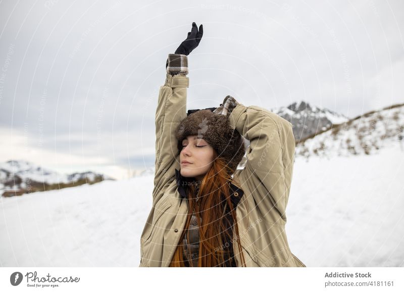 Dreamy traveler in outerwear against snowy mountain eyes closed breathe enjoy harmony idyllic winter woman arm raised ornament glove wellness trip journey