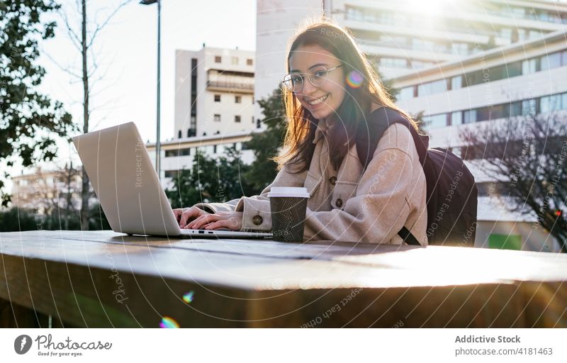 Cheerful female student working on laptop in city park woman toothy smile assignment cheerful using campus gadget break coffee backpack eyeglasses to go