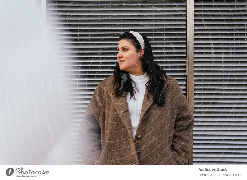 Dreamy plump woman in coat near wall in town hand in pocket contemplate casual style friendly enjoy sincere fence sky building cloudy idyllic spare time weekend