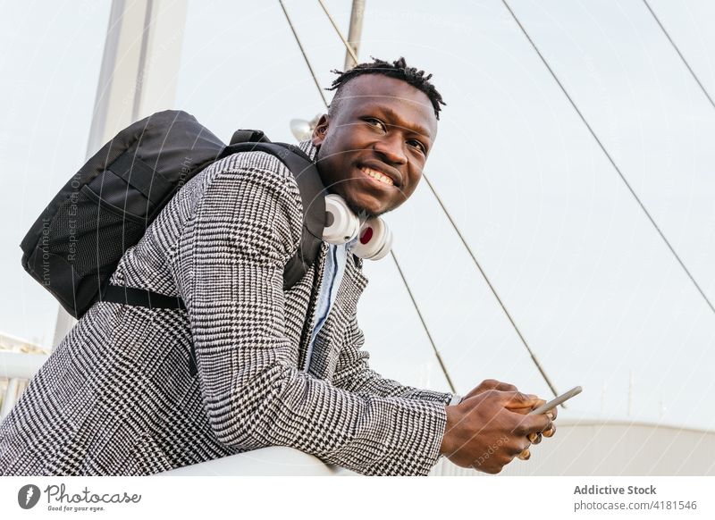 Glad black office worker with smartphone on suspension bridge manager smile headphones formal backpack lean forward town gadget device dreadlocks headset