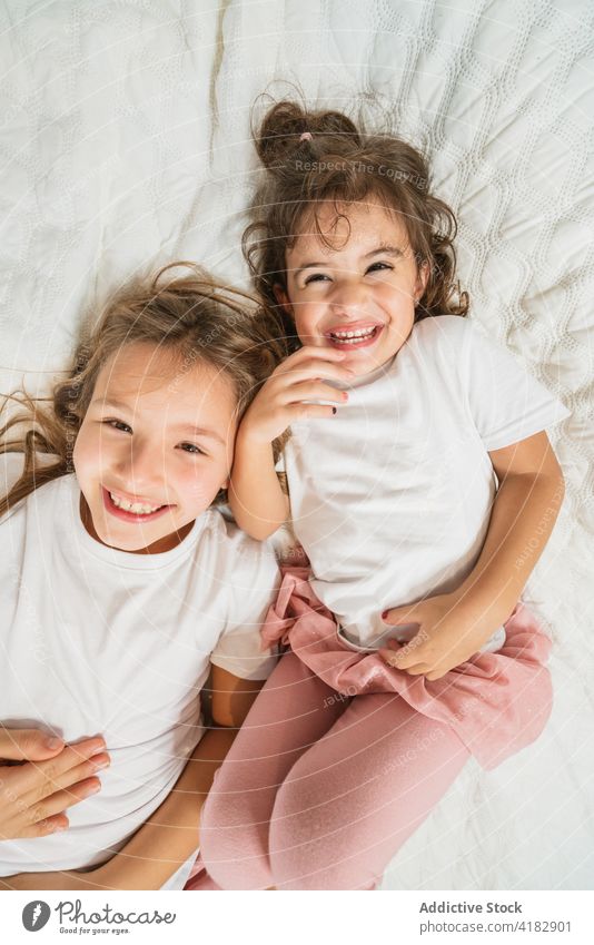 Joyful sisters lying on cozy bed looking at camera girl excited having fun joy cheerful happy sibling energy childhood kid casual carefree positive optimist