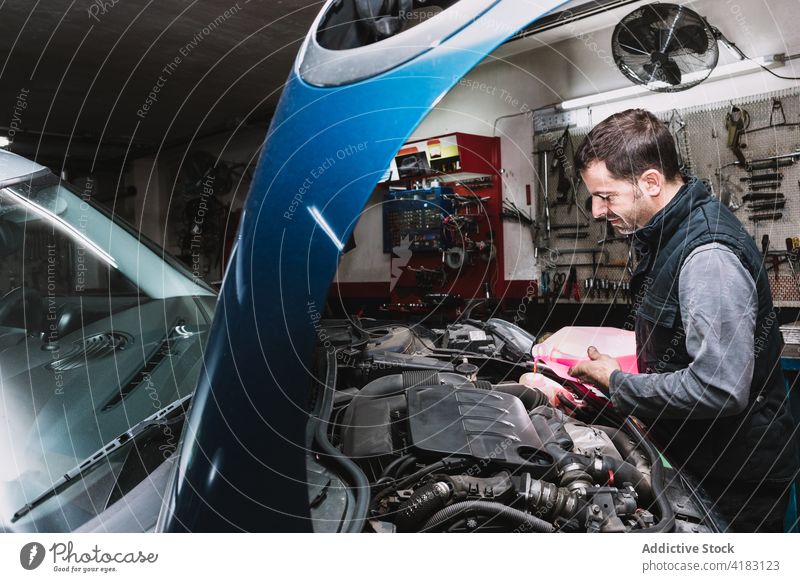 Smiling mechanic pouring coolant into radiator in auto workshop hood vehicle maintenance man garage bonnet mechanism hose wiper unshaven liquid plastic canister