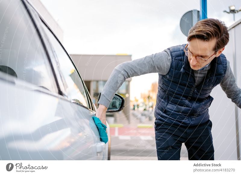 Concentrated young man wiping car with napkin wash wipe rag focus vehicle self service auto station street window water automobile manual male casual eyeglasses