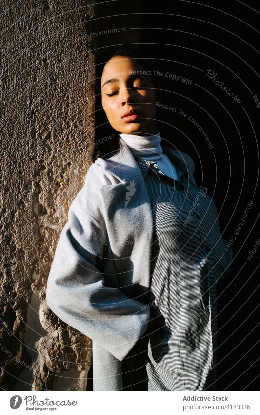 Stylish woman with eyes closed leaning against stone wall style sunlight serene tranquil relax calm fashion season young ethnic female coat trendy lifestyle