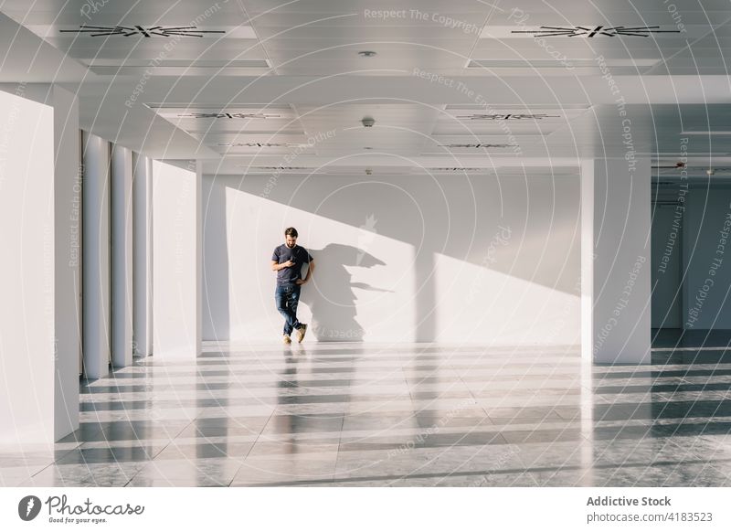 Man standing in empty spacious office hall and browsing smartphone man facility shadow using male interior sunlight geometry shape hallway white wall surfing